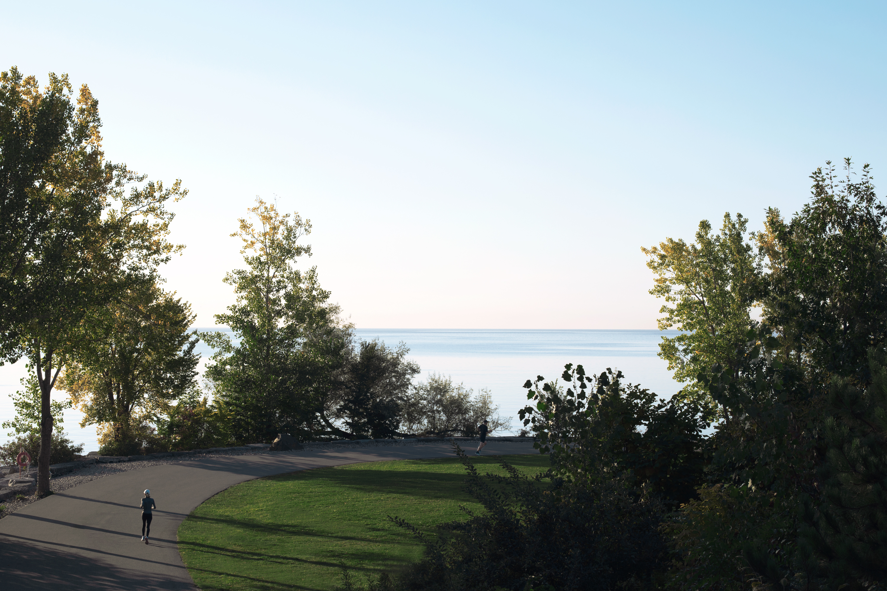 Runners on a waterfront trail lined with trees looking out over Lake Ontario at sunrise