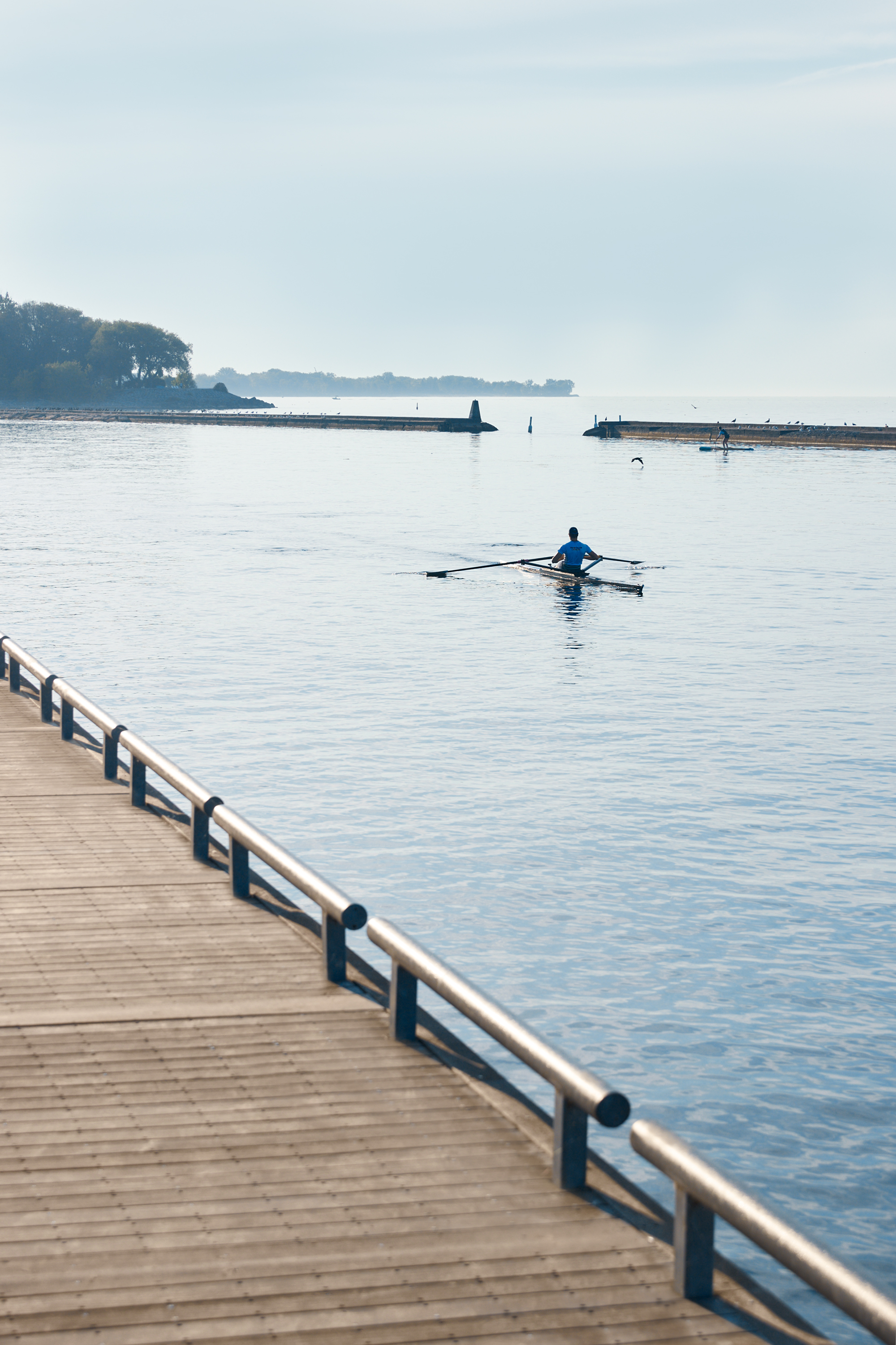 A rower and paddleboarder on calm lake water beside a boardwalk